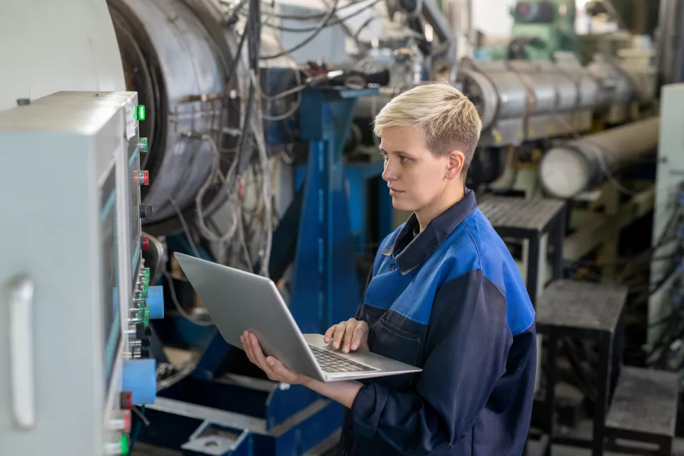 young-serious-blond-female-worker-overalls-using-laptop-while-looking-through-online-technical-data-against-industrial-equipment-workshop
