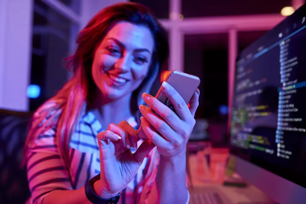 cheerful-female-coder-working-late-night-office-with-neon-light-texting-friends-focus-hands